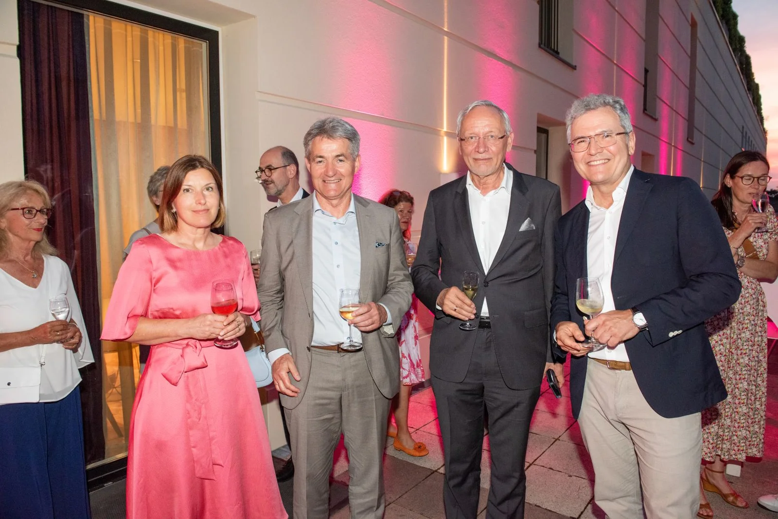 Eine Gruppe von Menschen steht draußen, formell gekleidet, bei einer Veranstaltung mit pinker Beleuchtung.