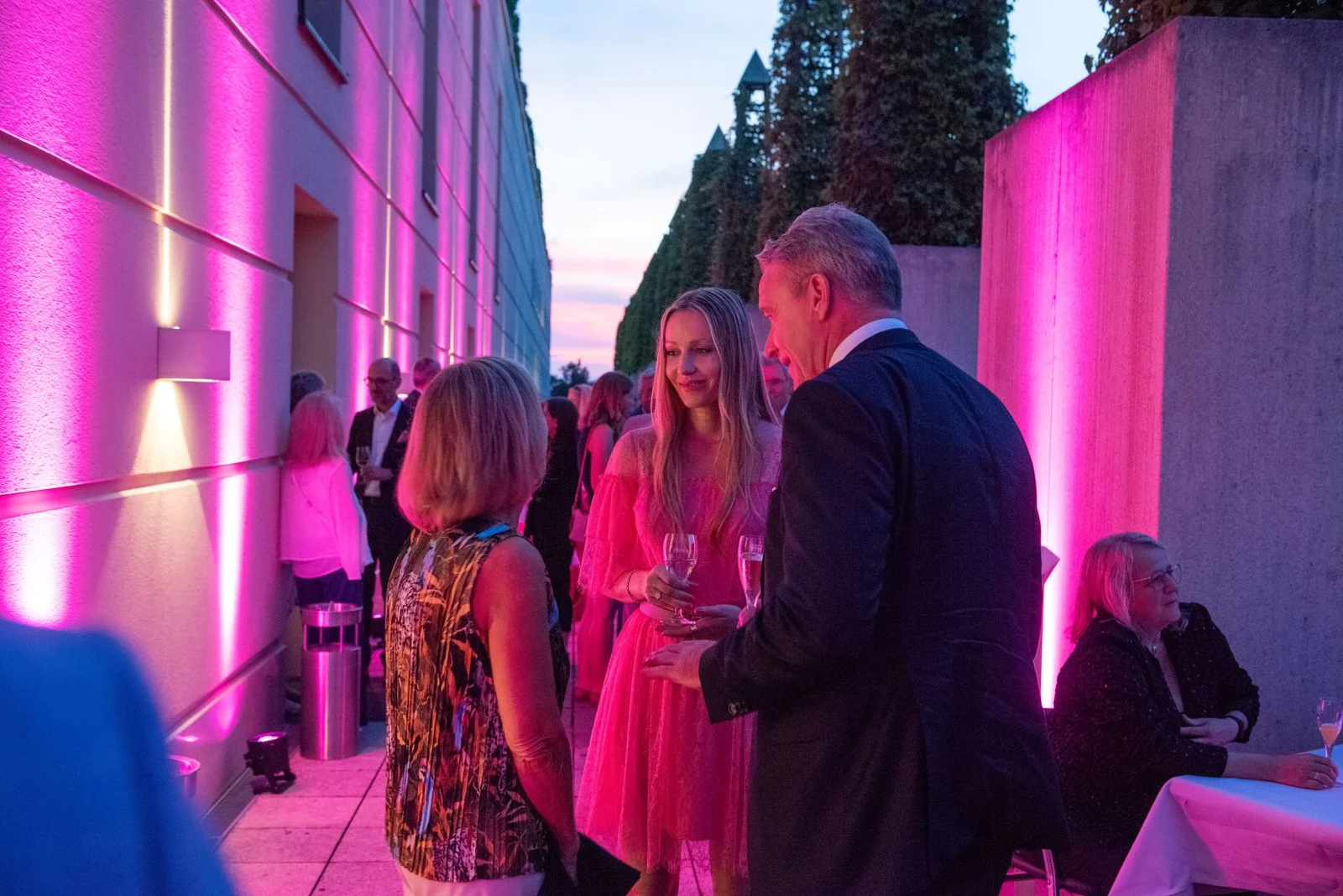Eine elegante Abendveranstaltung im Freien mit Menschen in formeller Kleidung. Pinke Lichter beleuchten die Szene.