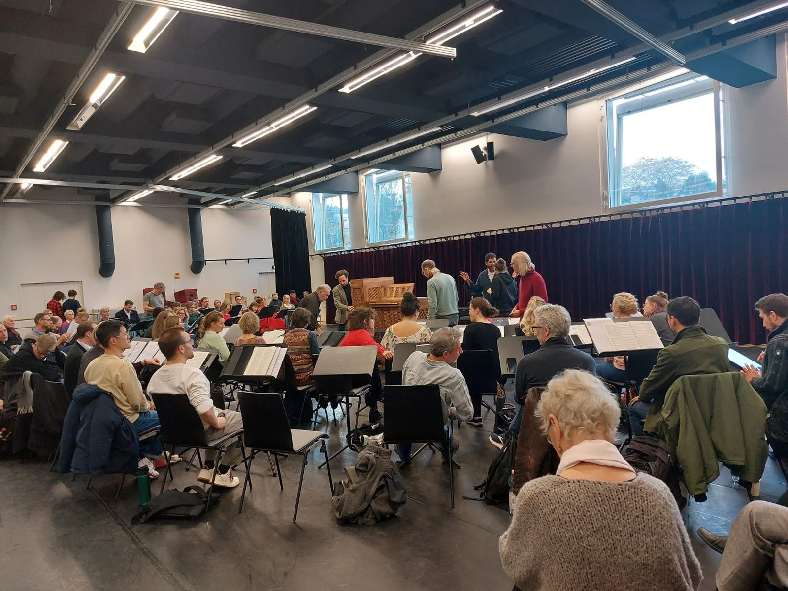 Ein großer Raum voller Menschen bei einer Orchesterprobe. Sitzplätze sind mit Notenpulten ausgestattet. Eine gemütliche, konzentrierte Atmosphäre.