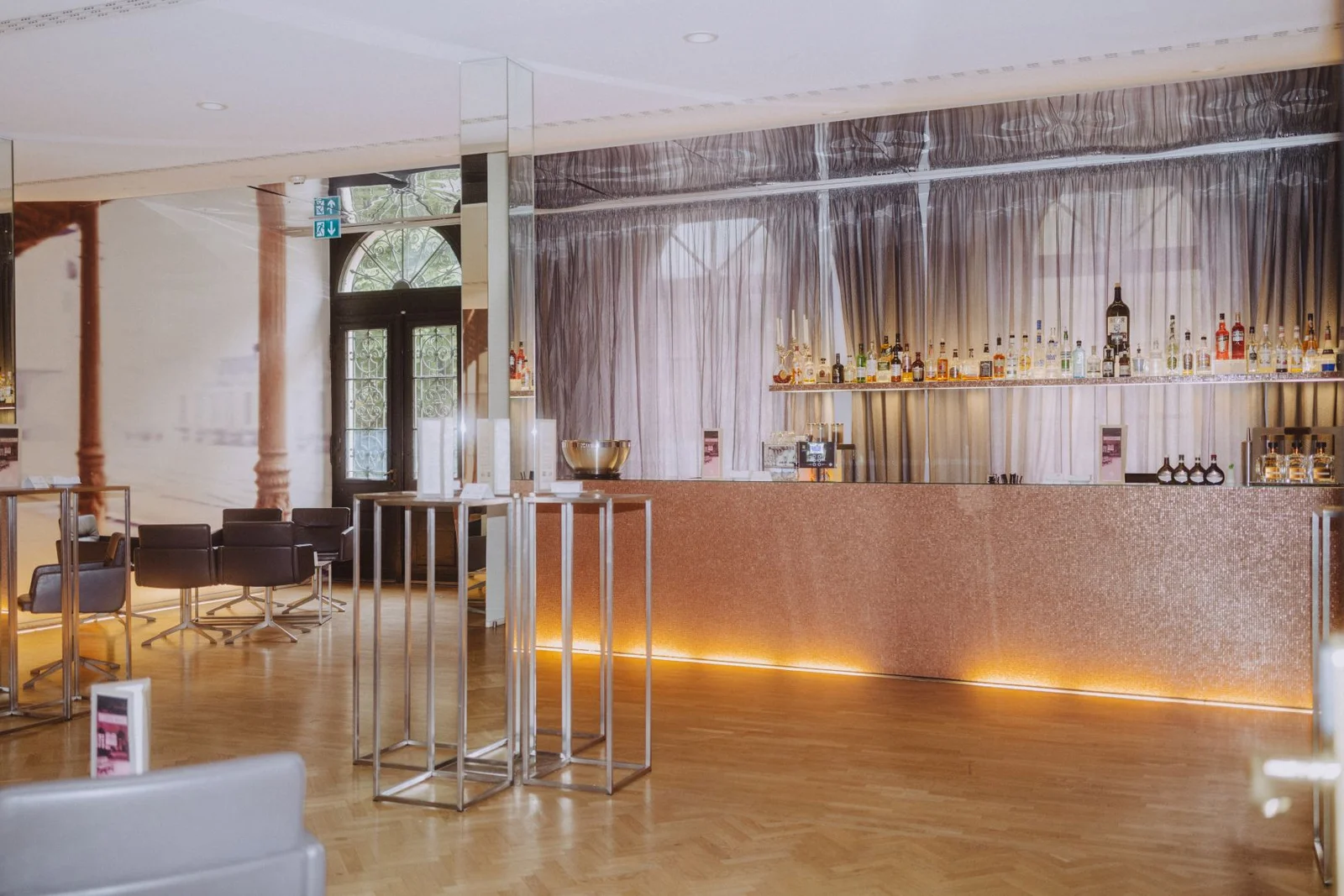 Moderne Bar mit eleganter Einrichtung, hohen Kerzenständern und einer Auswahl an Flaschen. Holzboden und große Fenster im Hintergrund.
