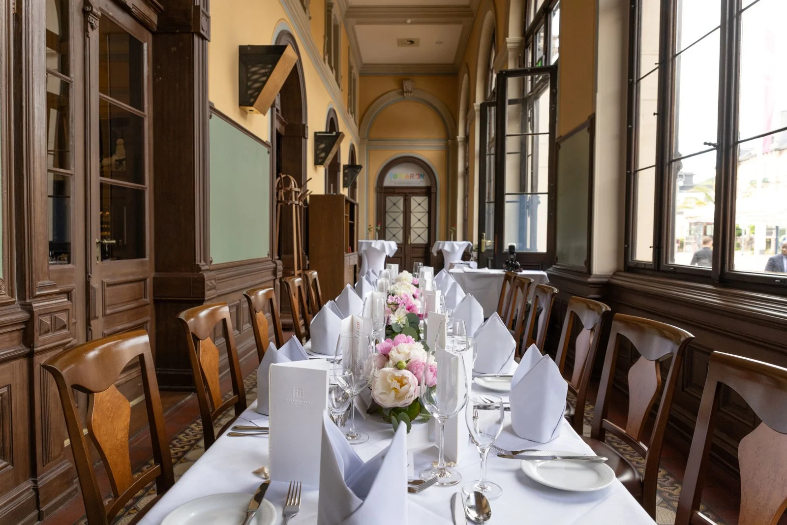 Elegant gedeckter langer Tisch in einem traditionellen Wintergarten mit Blumen in der Mitte und gefalteten Servietten.