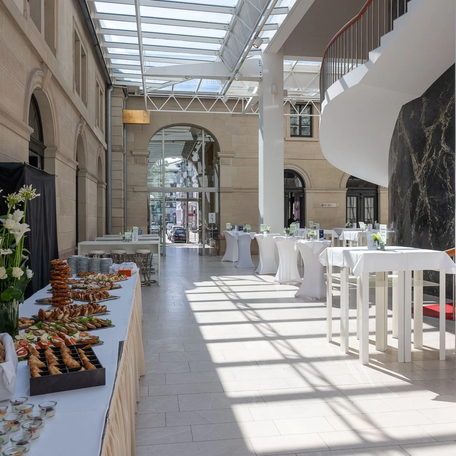 Ein eleganter Veranstaltungsraum mit hohen Decken und großen Fenstern. Weiße Stehtische und ein Buffet mit Snacks. Helle, einladende Atmosphäre.