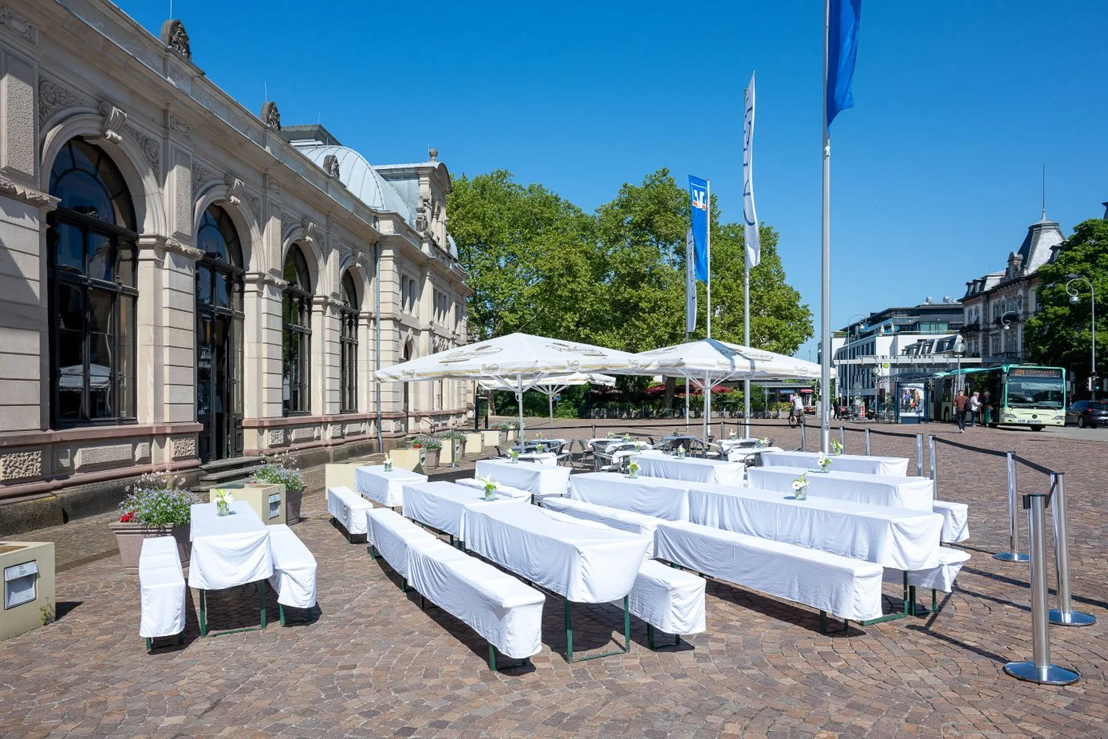 Weiße Tische und Bänke sind im Freien auf einem gepflasterten Platz neben dem Festspielhaus aufgestellt. Sonnenschirme spenden Schatten.