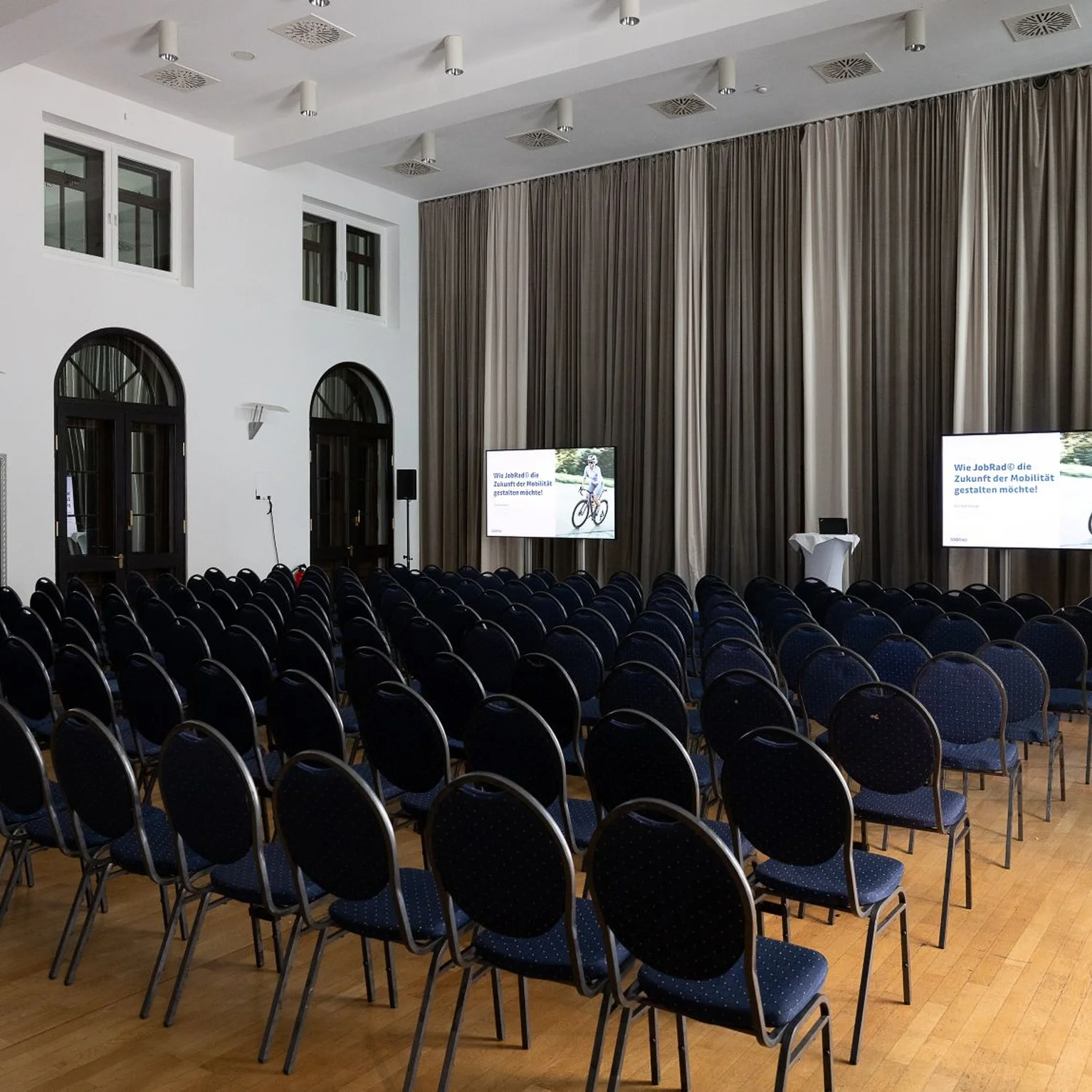 Ein leerer Konferenzraum mit vielen schwarzen Stühlen, zwei Bildschirmen vorne und großen Fenstern an den Wänden.