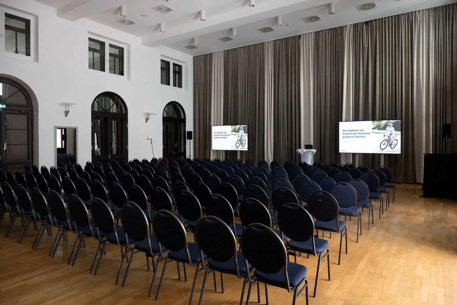 Ein leerer Konferenzraum mit vielen schwarzen Stühlen, zwei Bildschirmen vorne und großen Fenstern an den Wänden.