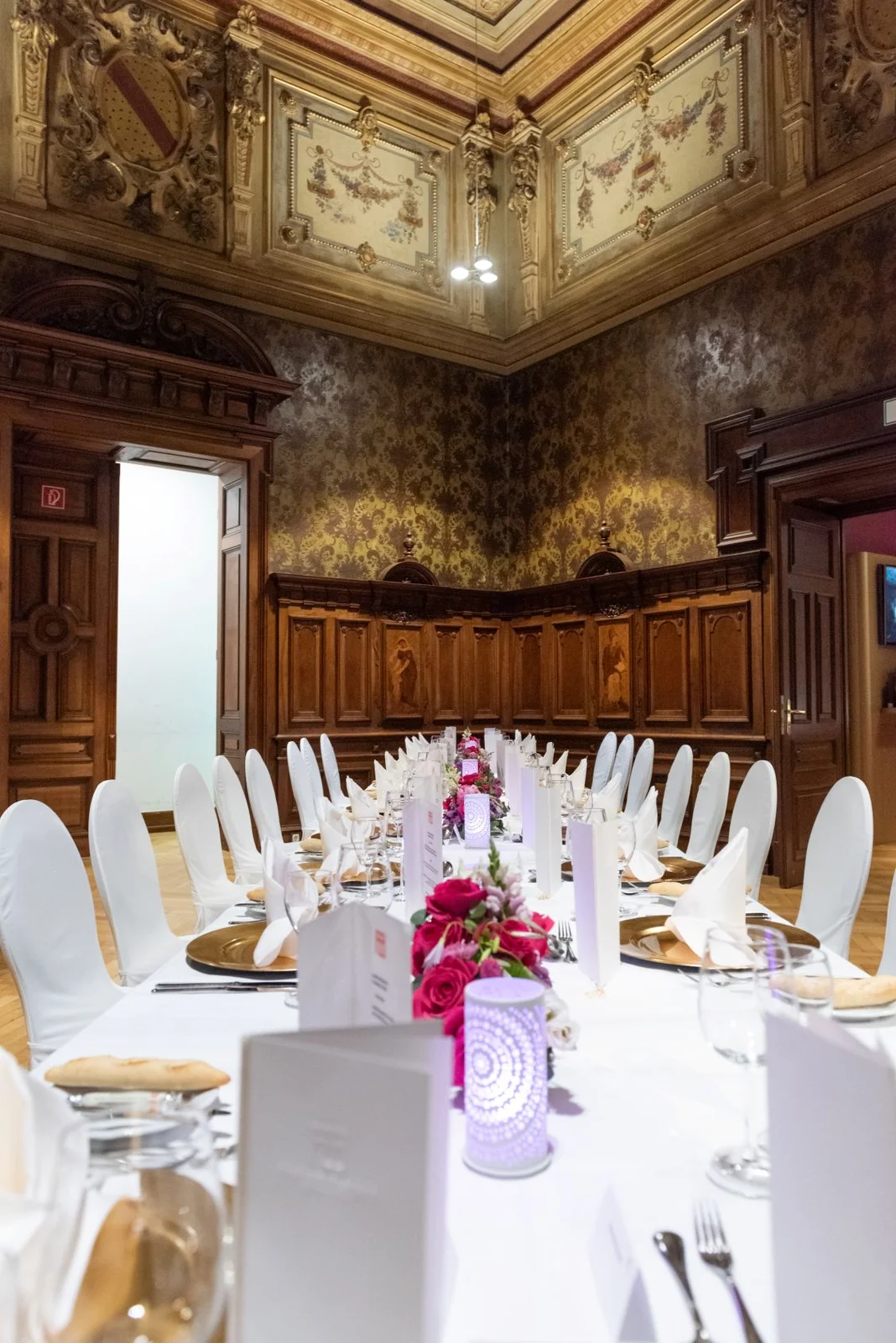 Elegante Tafel in einem opulenten Raum mit Holztäfelung und reich verziertem Deckengemälde. Weiß gedeckte Tische mit rosa Blumendekorationen.