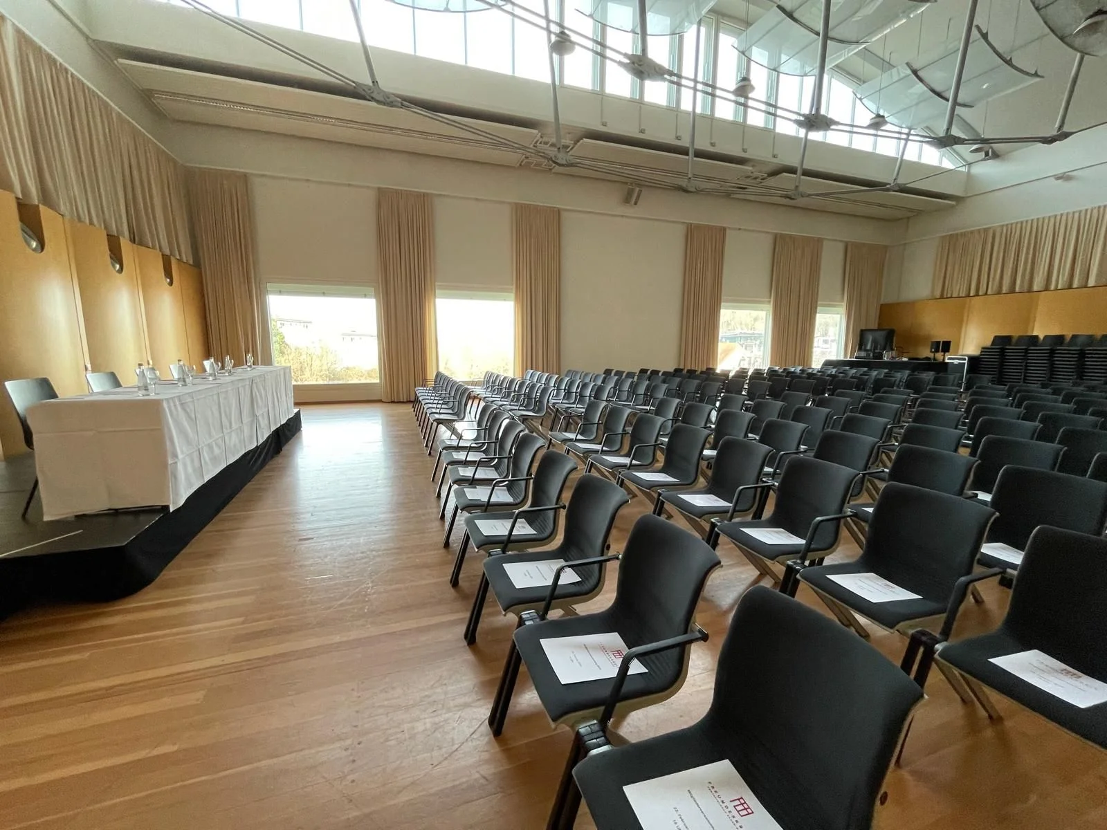 Der leere Orchesterprobensaal mit Reihen schwarzer Stühle und einem Tisch vorne. Große Fenster lassen viel Licht herein.
