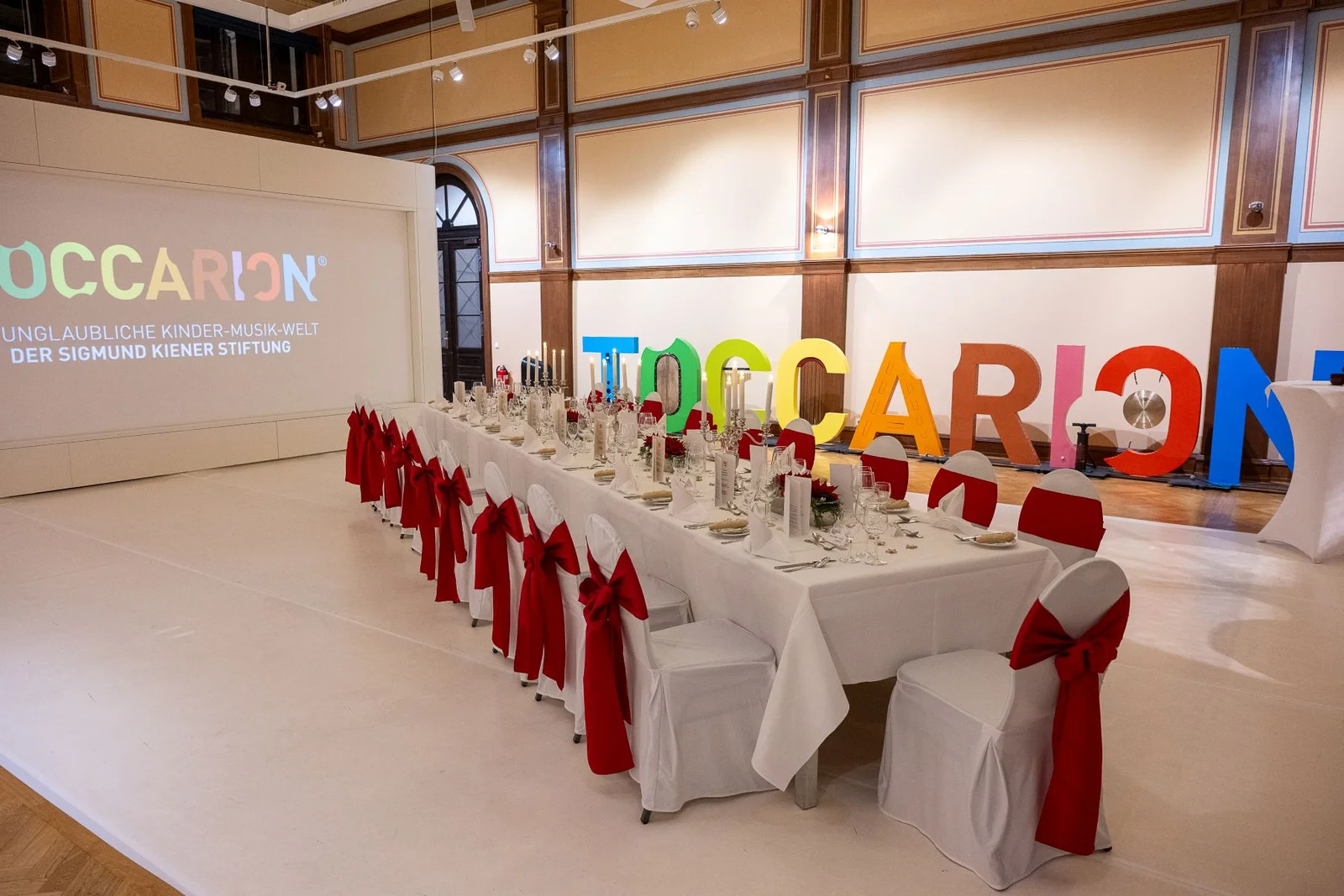 Ein elegant gedeckter Tisch mit roten Stuhlschleifen in einem Raum mit großen, bunten TOCCARION-Buchstaben und einer Projektionsleinwand.