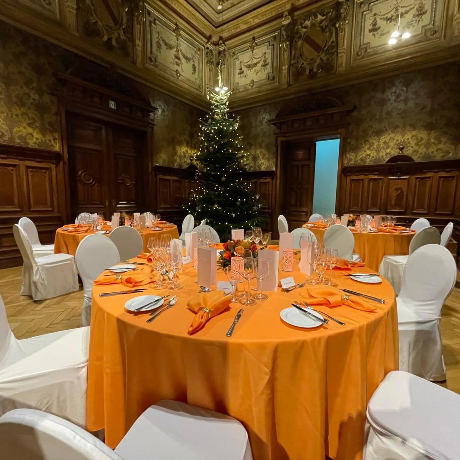 Eleganter Raum mit gedeckten Tischen und orangefarbenen Tischdecken, weißen Stühlen und einem festlich geschmückten Weihnachtsbaum im Hintergrund.