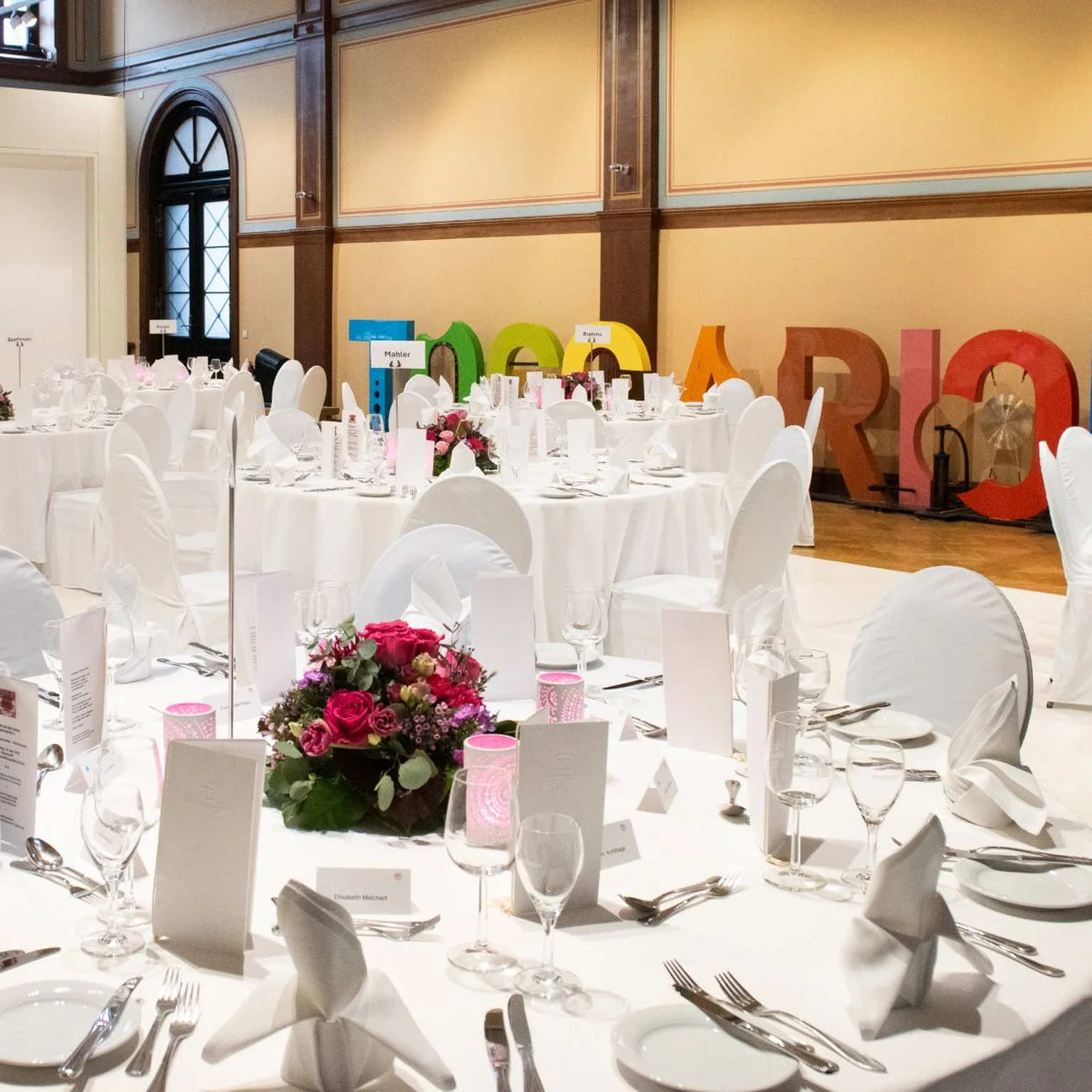 Ein eleganter Veranstaltungsraum mit runden Tischen, weiß gedeckten Stühlen und Blumenarrangements. Bunte Buchstaben an der Wand formen das Wort "Toccarion".