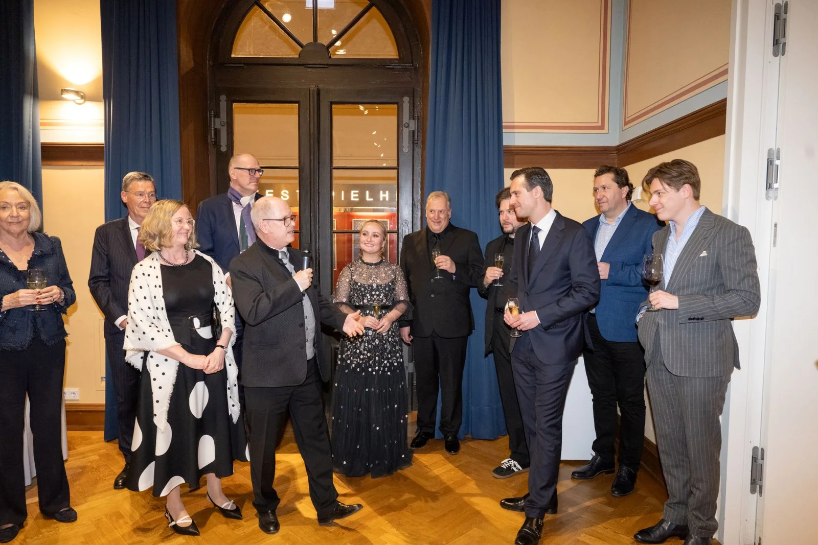 Eine Gruppe elegant gekleideter Menschen steht in einem Raum und unterhält sich. Der Raum hat Holzböden und blaue Vorhänge.