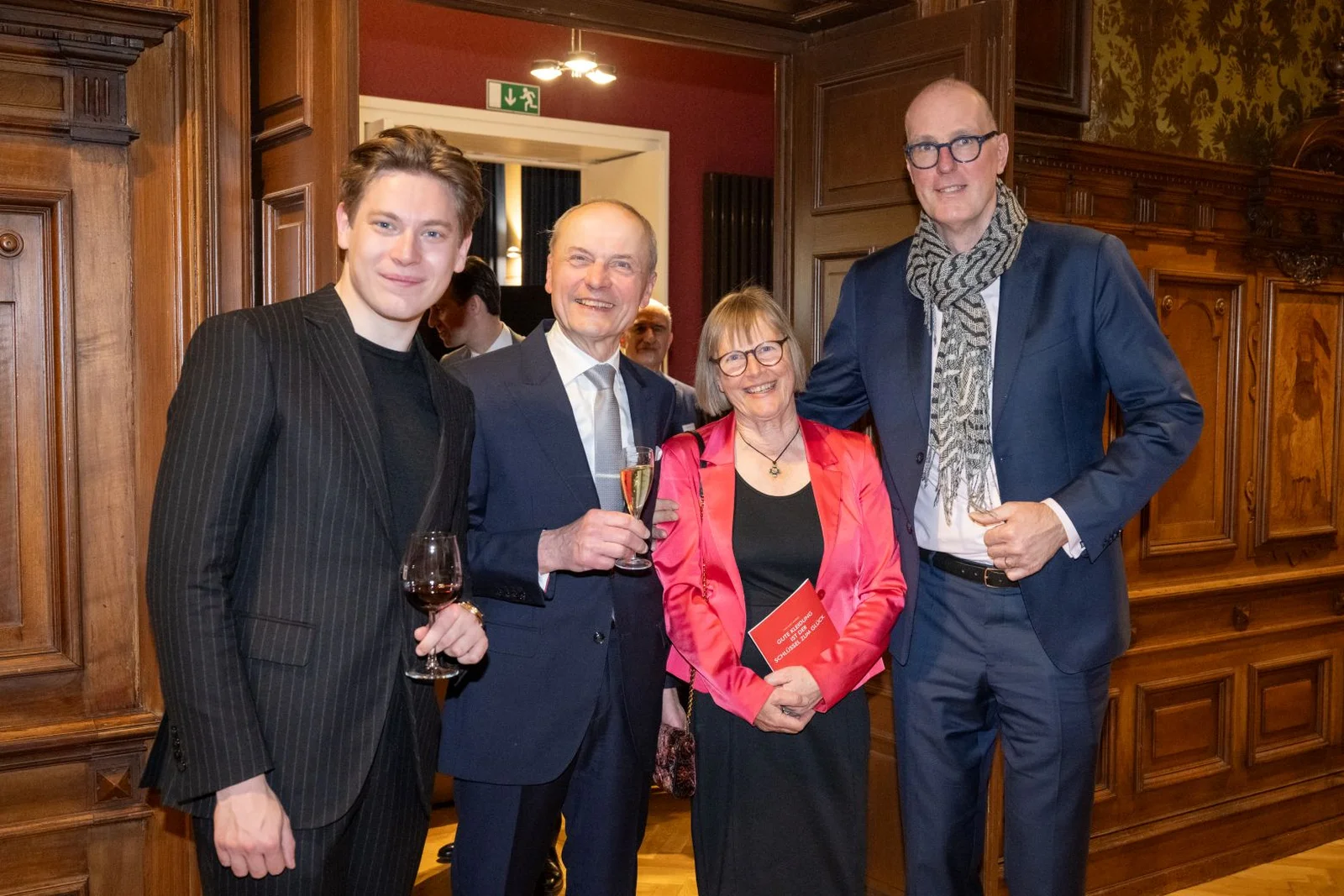 Vier Personen stehen in einem eleganten Raum, halten Getränke und lächeln in die Kamera. Die Atmosphäre wirkt freundlich und gesellig.