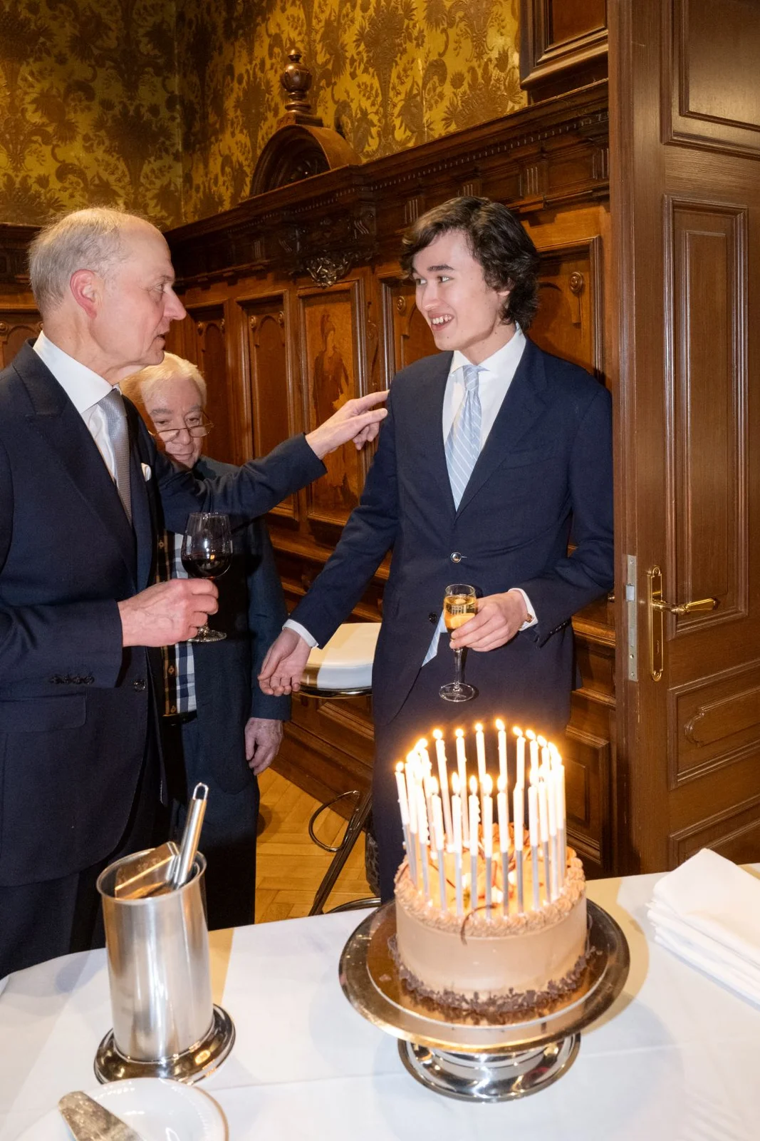 Drei Männer in Anzügen befinden sich in einem eleganten Raum mit einer Torte voller Kerzen auf einem Tisch.