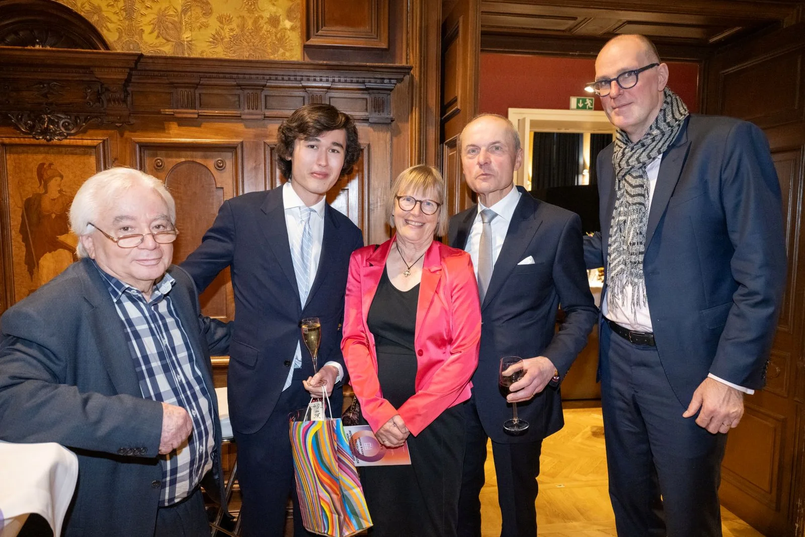 Fünf Personen posieren in formeller Kleidung für ein Gruppenfoto in einem eleganten Raum. Zwei halten Getränke in der Hand.
