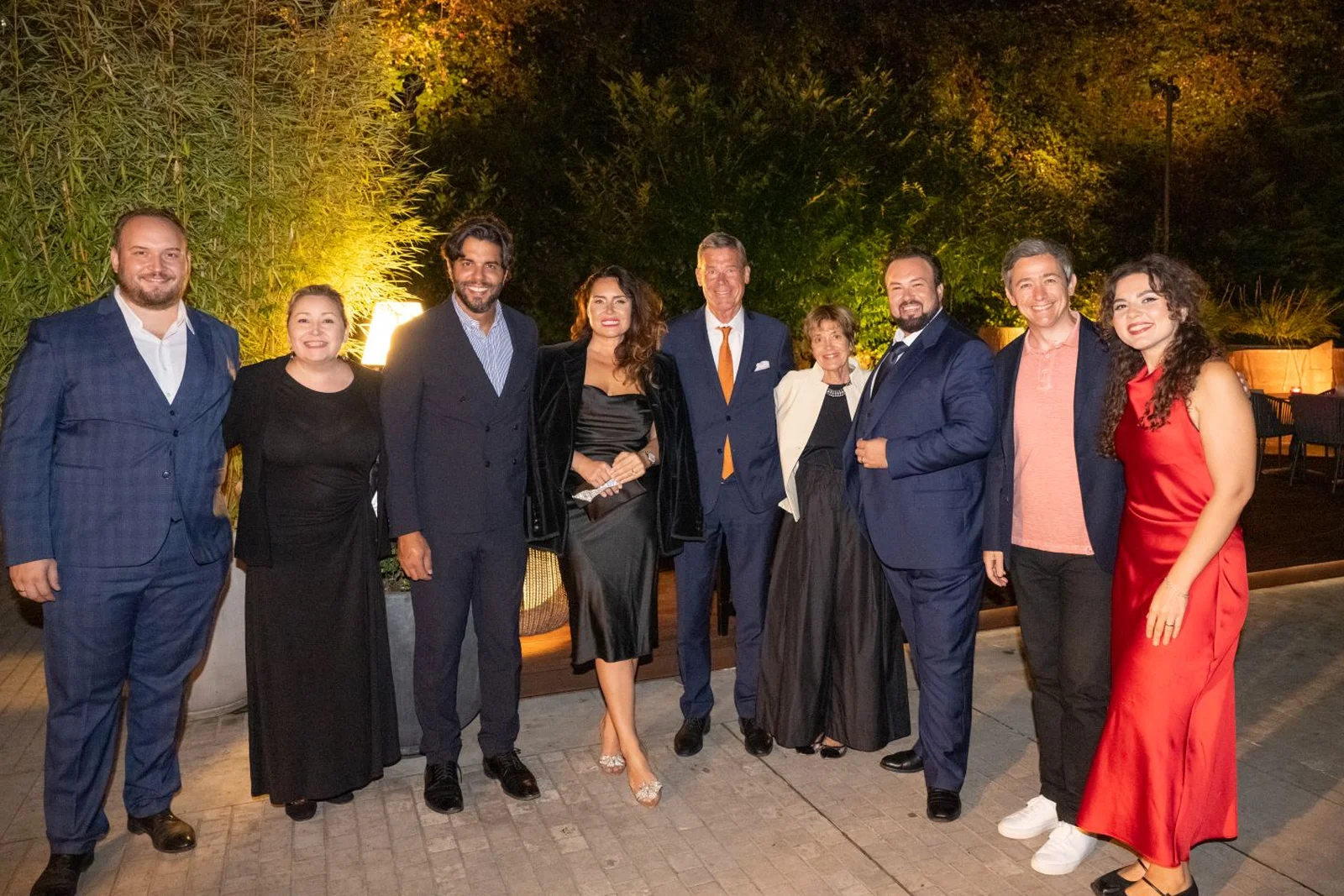 Eine Gruppe eleganter Menschen posiert nachts im Freien für ein Foto. Sie tragen formelle Kleidung und lächeln fröhlich.