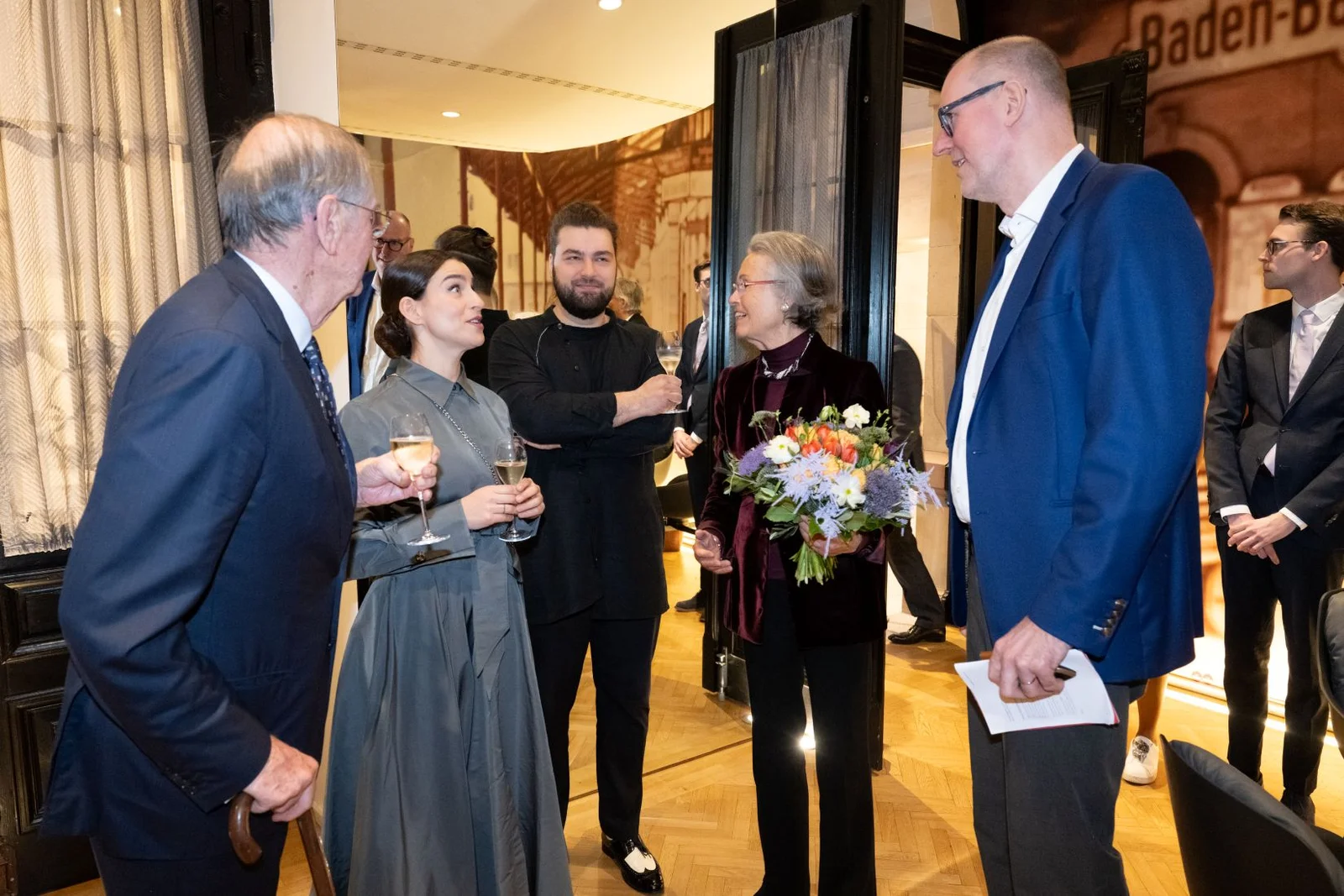 Mehrere Personen stehen in einem eleganten Raum, eine Person hält Blumen. Sie unterhalten sich und trinken Getränke.