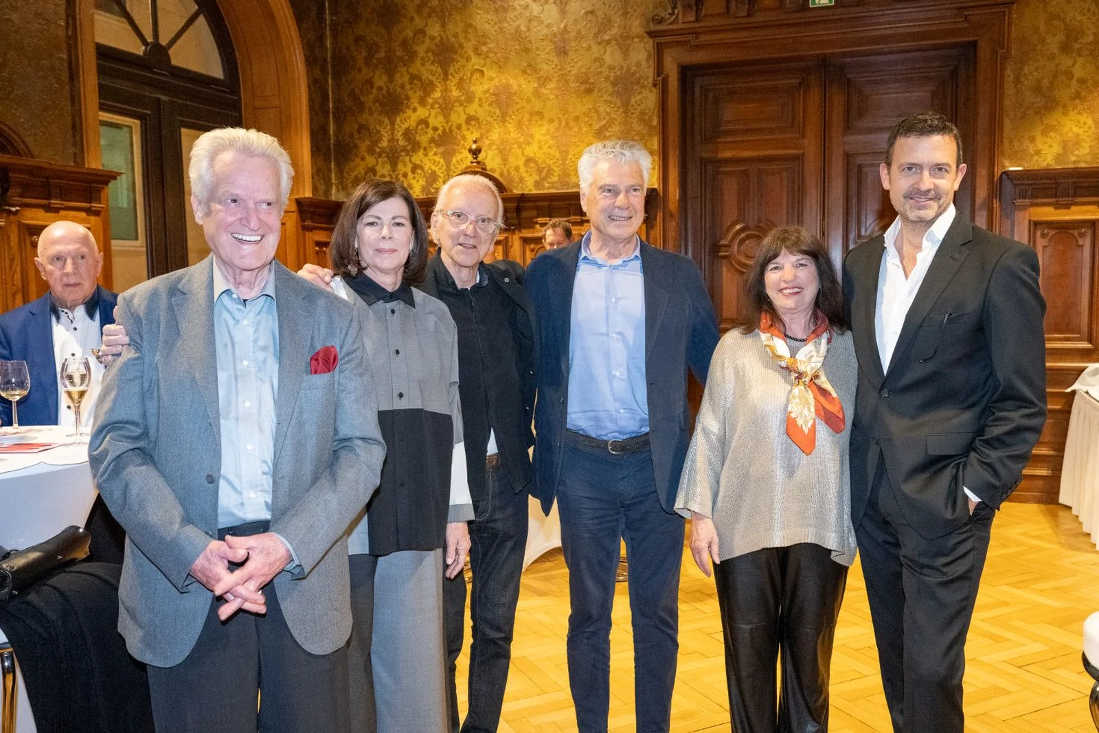 Eine Gruppe von sechs Menschen posiert lächelnd in einem eleganten Raum mit Holztäfelungen und Tischen im Hintergrund. Sie tragen formelle Kleidung.