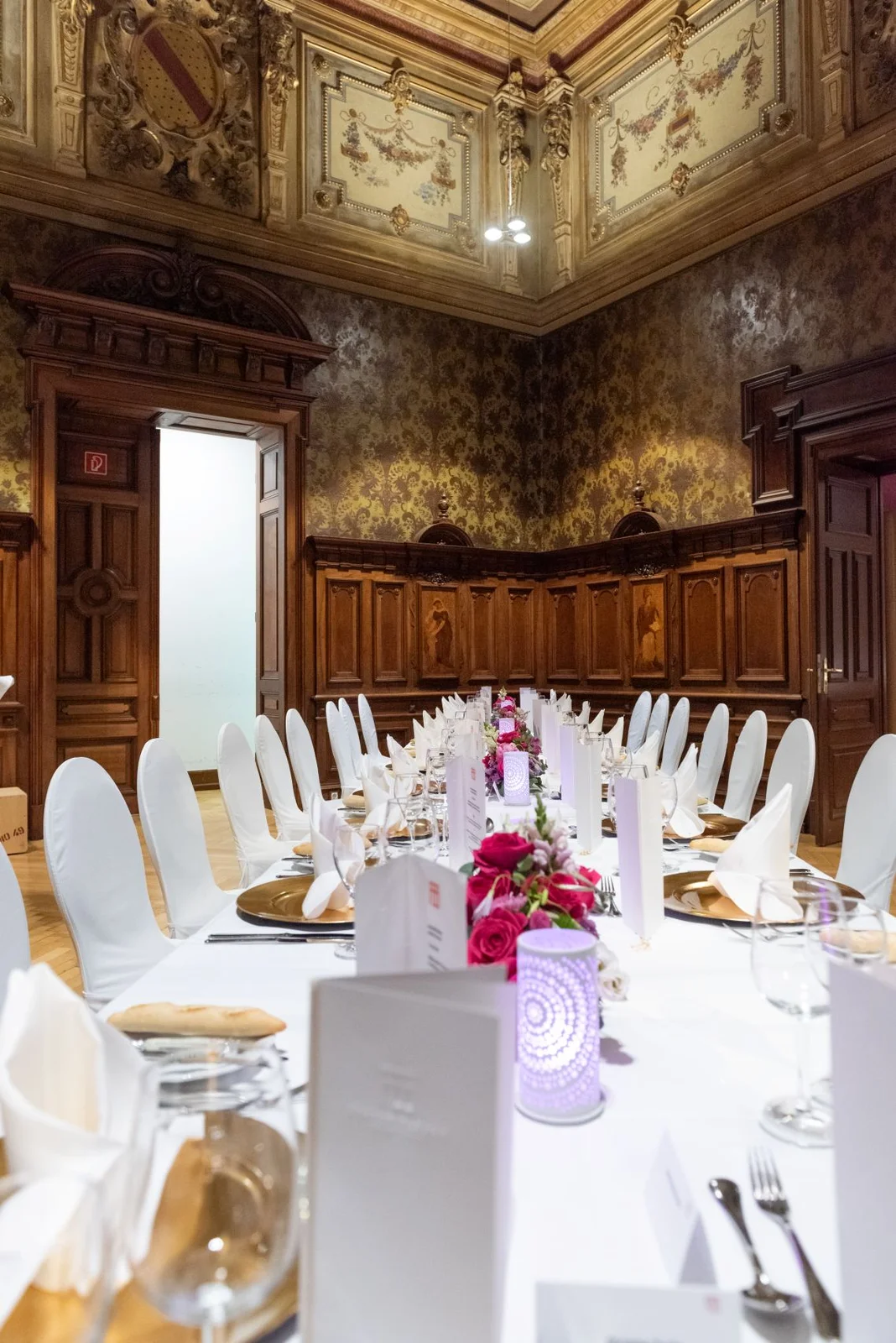 Ein eleganter, reich verzierter Raum mit langen Tischen, weißen Stuhlhussen und dekorativen Blumen.