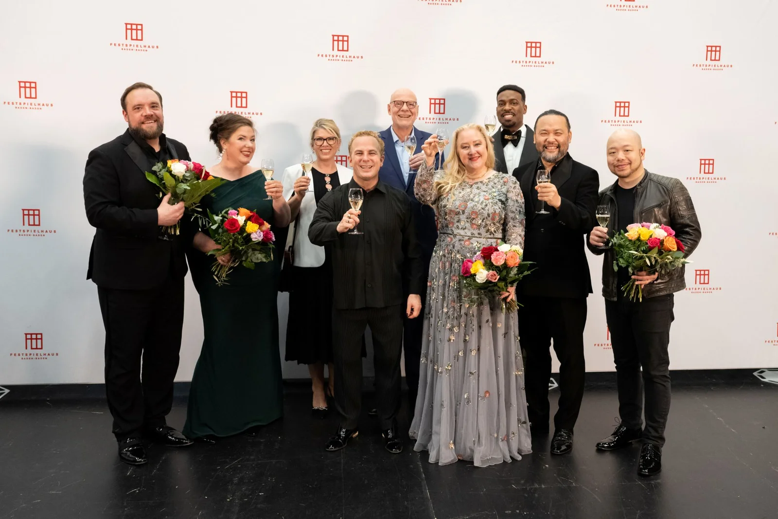 Eine Gruppe von zehn Personen in Abendgarderobe posiert vor einem Hintergrund mit Logos. Einige halten Blumensträuße oder Gläser.