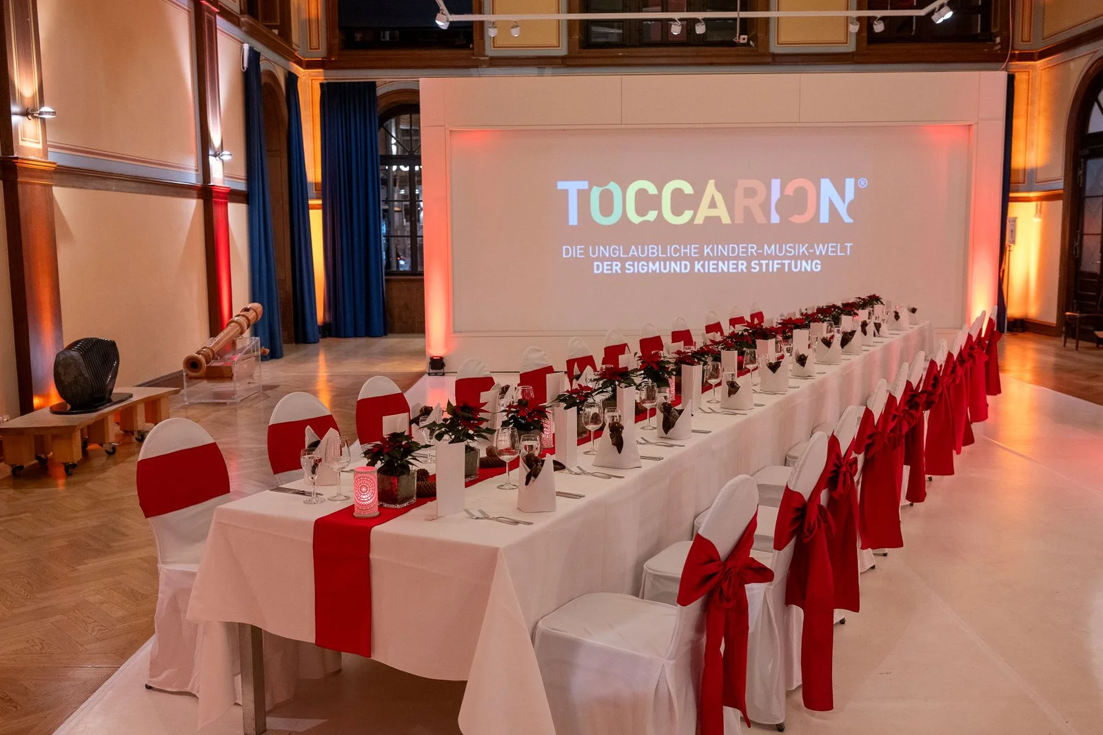 Ein eleganter Veranstaltungssaal mit weiß-roter Tischdekoration und Stühlen. Der Hintergrund zeigt das Wort "TOCCARION" auf einem Bildschirm.