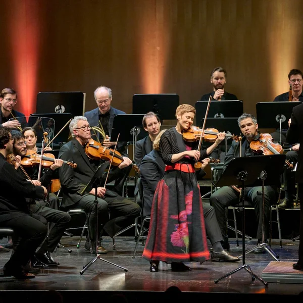 Ein Orchester spielt auf einer Bühne. Eine Frau in traditioneller Kleidung steht im Vordergrund und spielt Geige.
