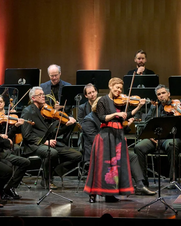 Ein Orchester spielt auf einer Bühne. Eine Frau in traditioneller Kleidung steht im Vordergrund und spielt Geige.