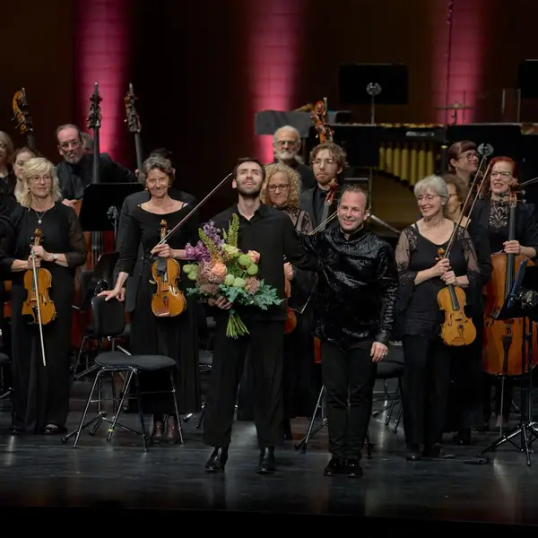 Der Dirigent und der Solist bedanken sich beim Publikum nach einem erfolgreichen Konzert. Die haben Blumensträuße in den Händen.