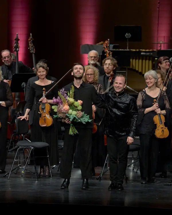 Der Dirigent und der Solist bedanken sich beim Publikum nach einem erfolgreichen Konzert. Die haben Blumensträuße in den Händen.