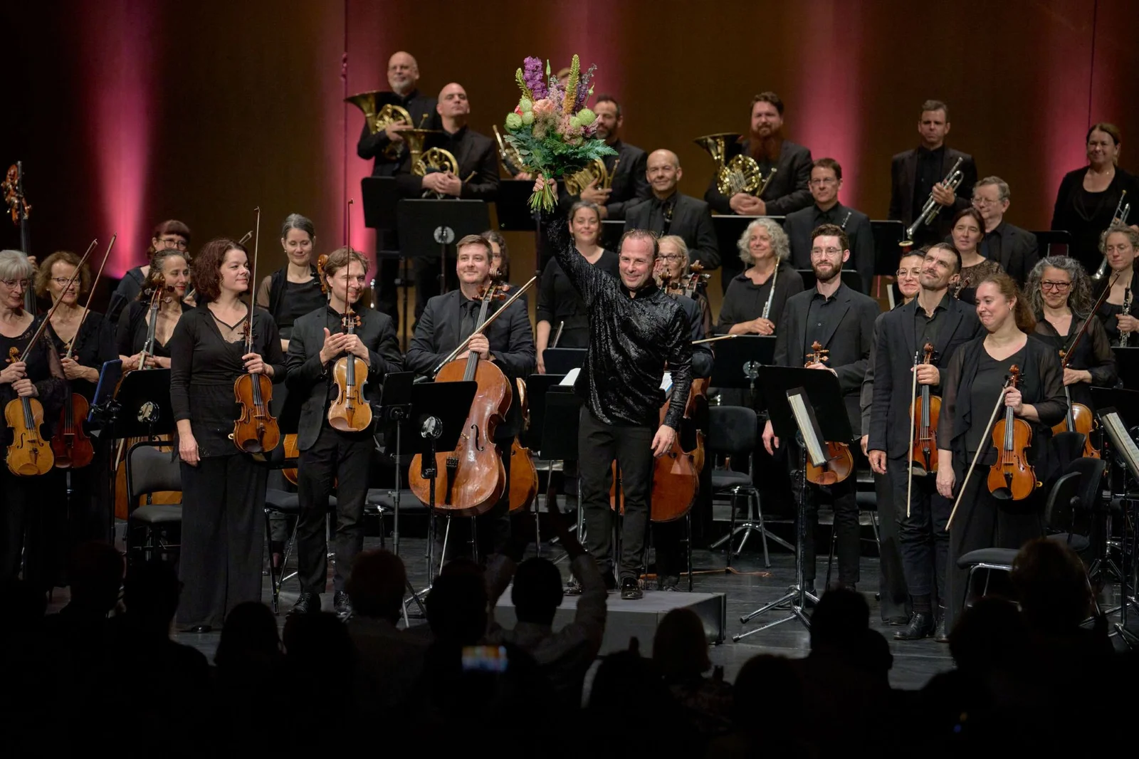 Ein Orchester steht auf einer Bühne. Der Dirigent hält einen Blumenstrauß. Musiker applaudieren vor einem Publikum.