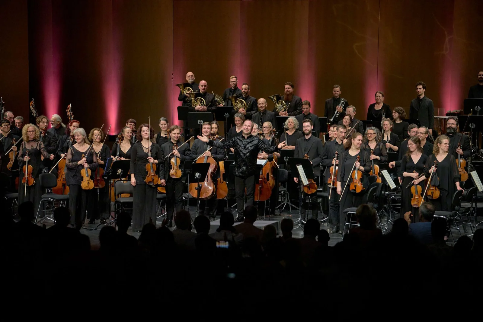 Ein Orchester auf einer Bühne, die Musiker tragen schwarze Kleidung und halten ihre Instrumente. Ein dunkler Hintergrund mit Beleuchtung.
