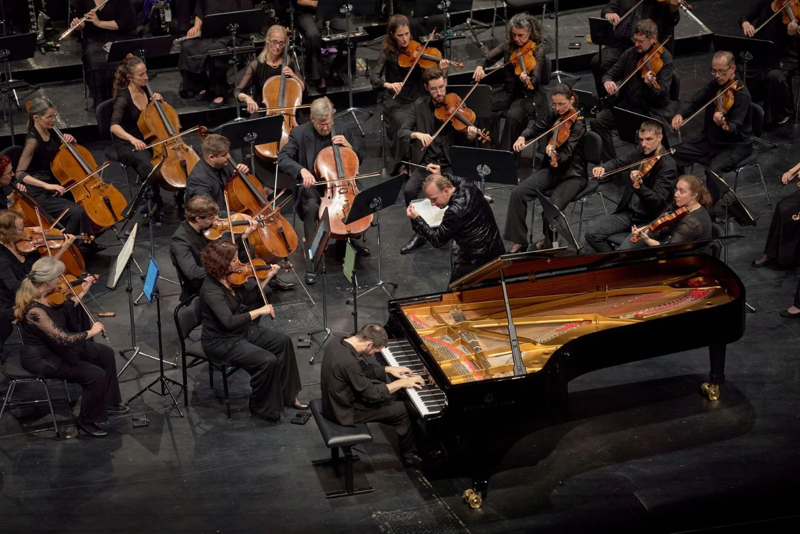 Ein Konzertflügel auf der Bühne, umgeben von einem Orchester mit Streichinstrumenten. Der Dirigent steht in der Mitte.