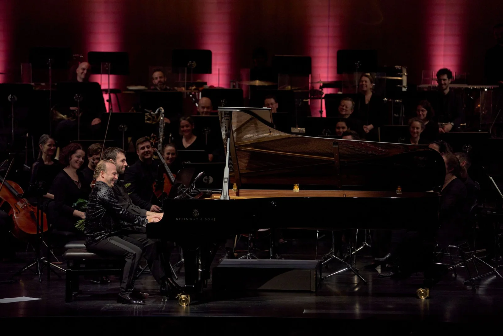 Ein Pianist spielt auf einem Konzertflügel, umgeben von einem Orchester. Der Hintergrund ist in rosa Licht getaucht.