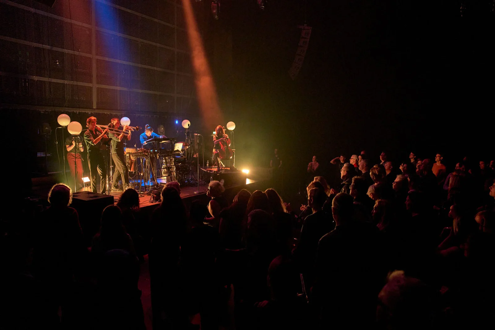 Ensemble spielt auf einer kleineren Bühne vor Publikum und wird mit orangenem atmosphärischen Licht angeleuchtet.