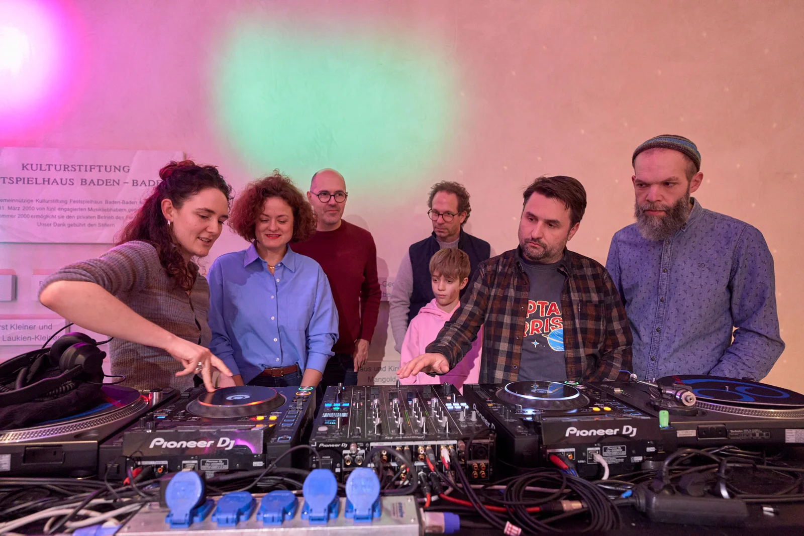 Eine Gruppe Menschen stehen hinter einem DJ-Pult, während der DJ die vielen Knöpfe und Regler erklärt.
