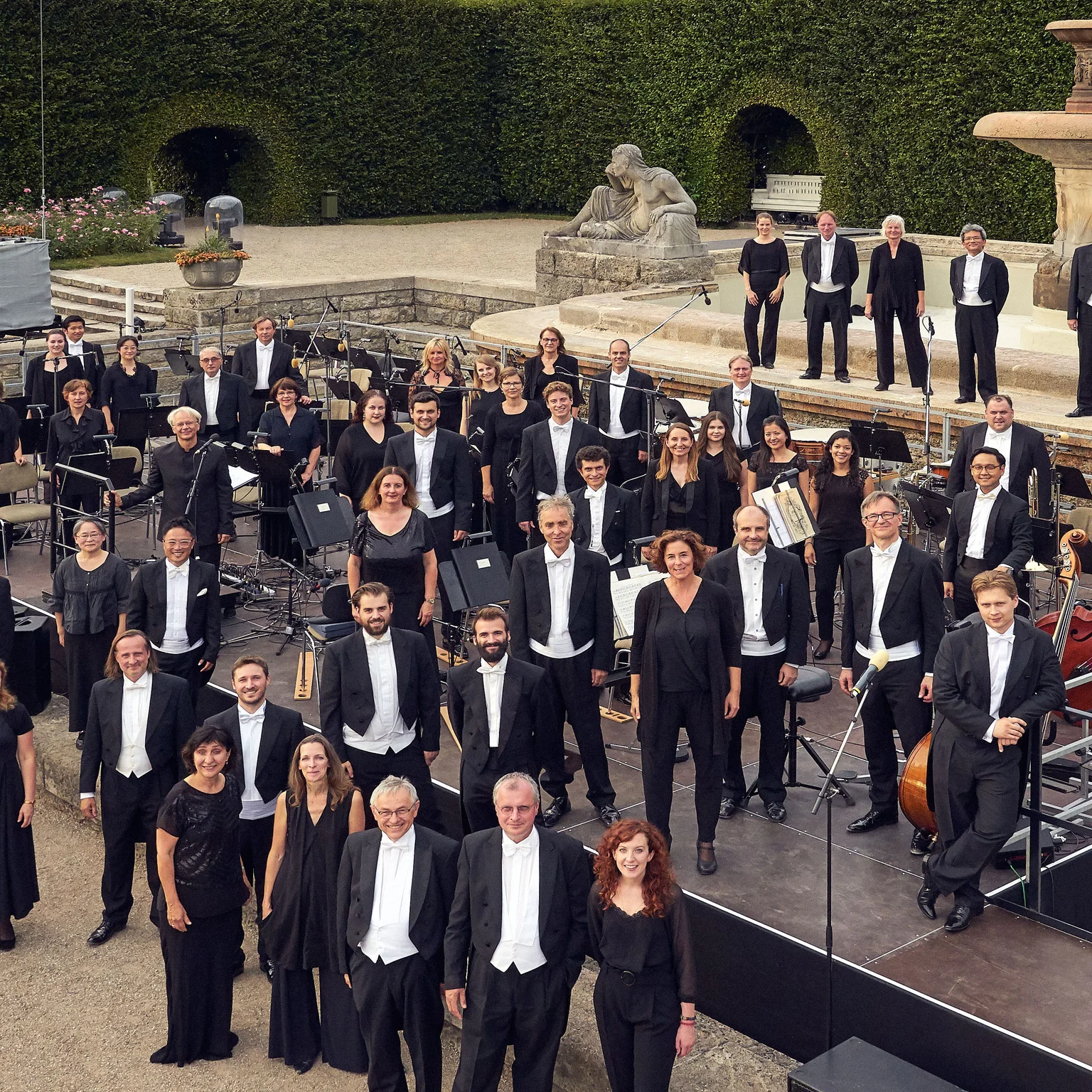 Ein großes Orchester posiert in formeller Kleidung, im Freien vor einer Brunnenanlage und dekorativen Hecken.