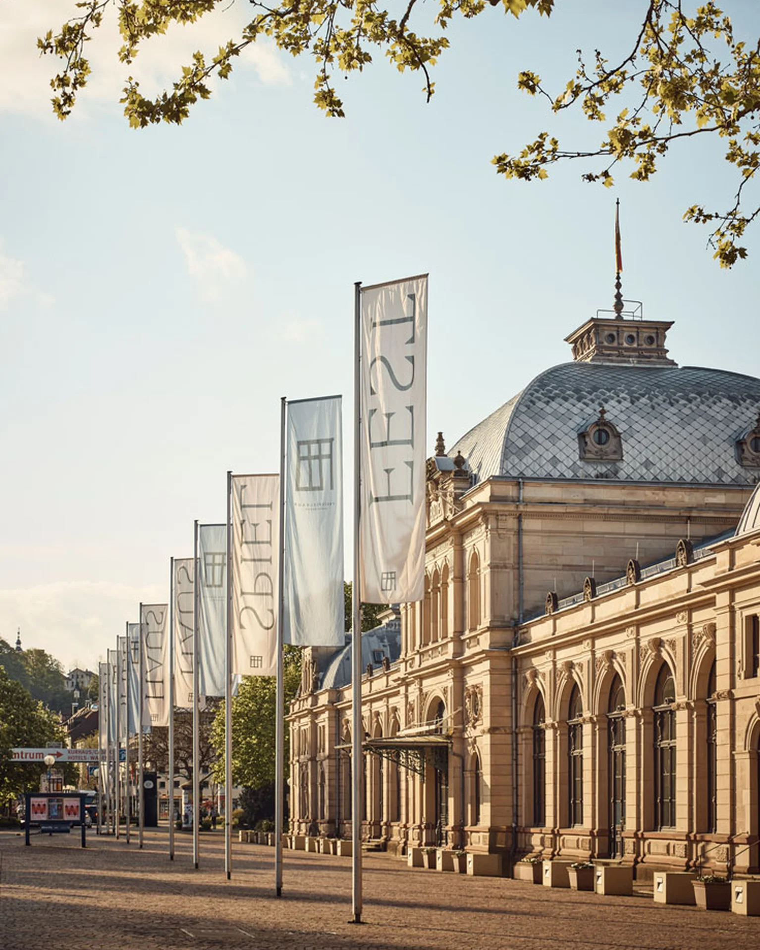 Festspielhaus Baden-Baden: Historisches Gebäude mit hoher Fassade und großen Fenstern. Drei lange Fahnen wehen vor dem Gebäude. Sonnenlicht beleuchtet die Szenerie.