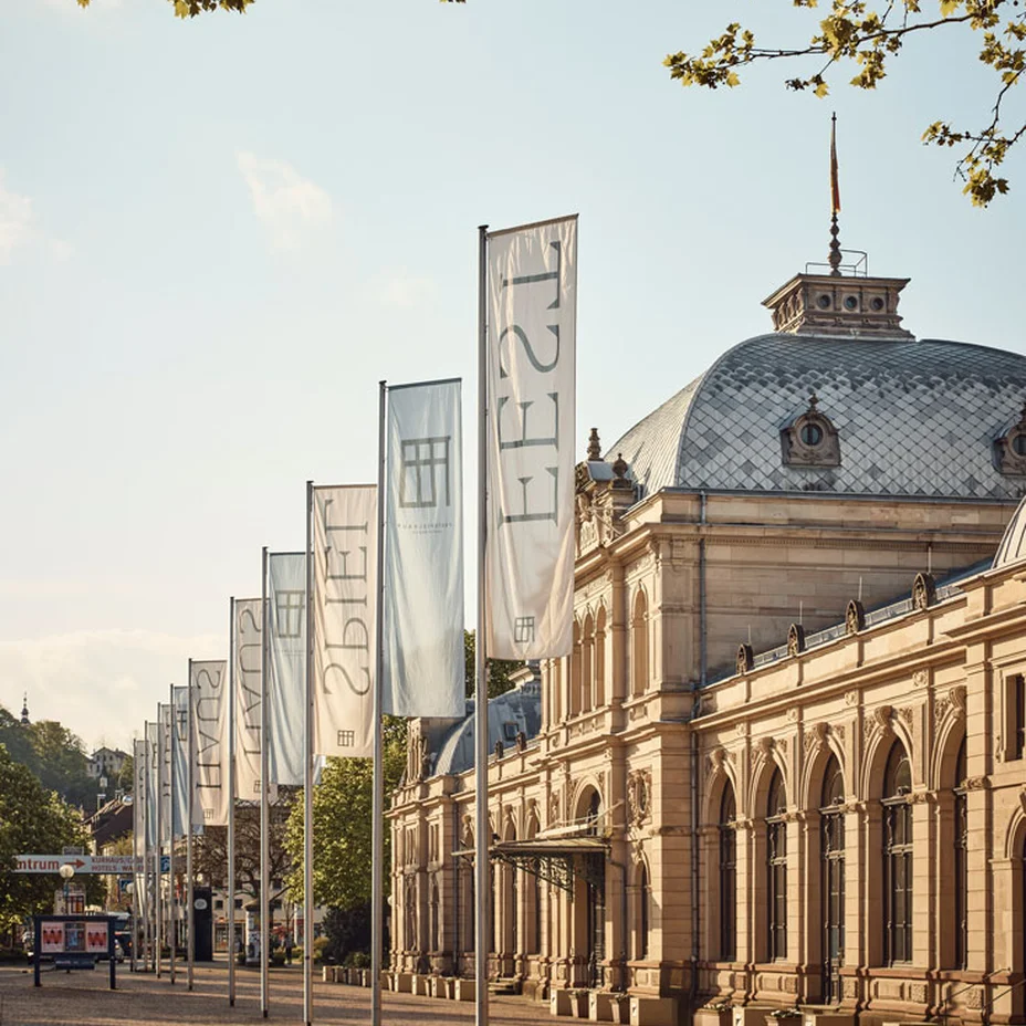 Festspielhaus Baden-Baden: Historisches Gebäude mit hoher Fassade und großen Fenstern. Drei lange Fahnen wehen vor dem Gebäude. Sonnenlicht beleuchtet die Szenerie.