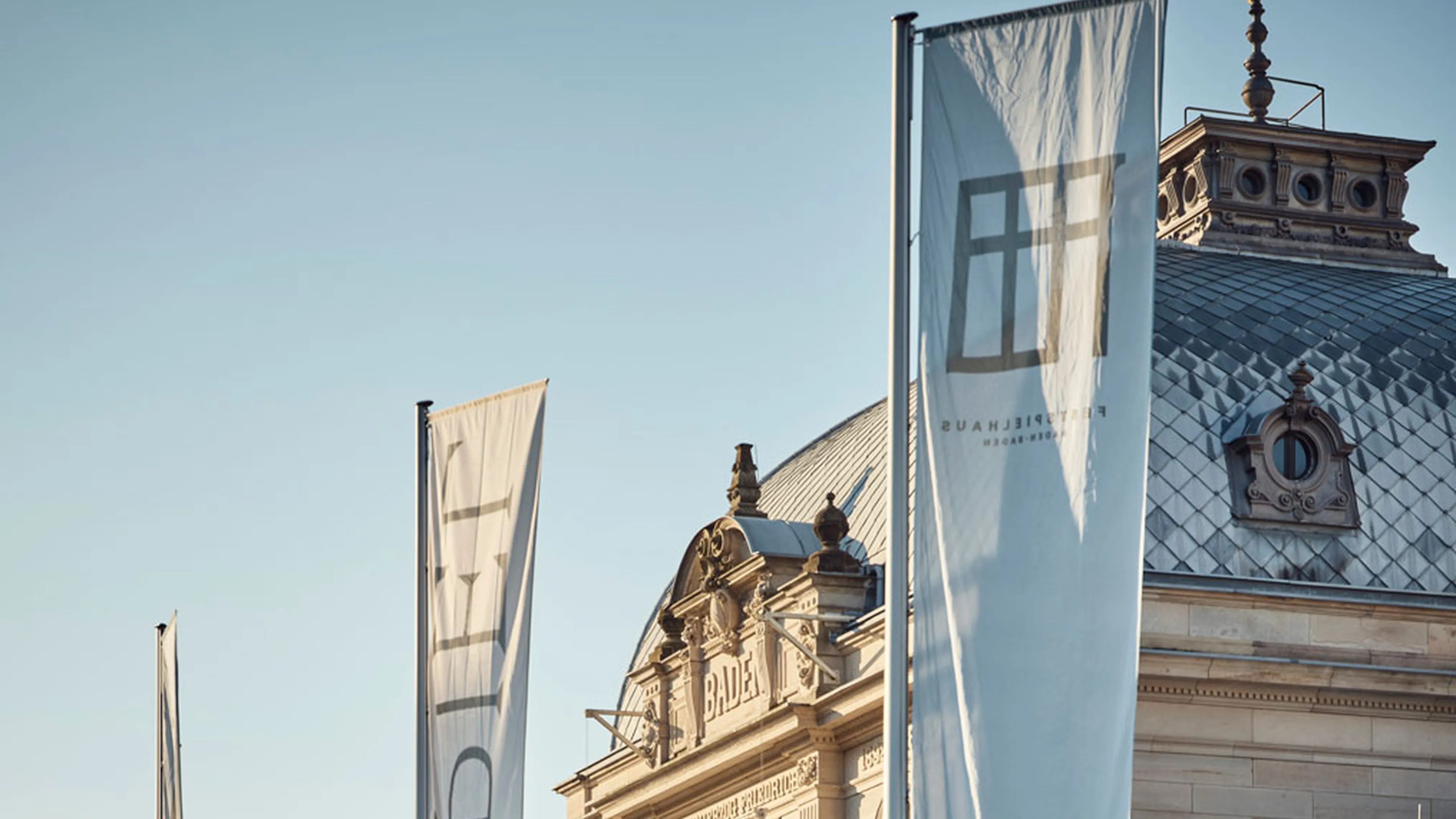 Festspielhaus Baden-Baden: Historisches Gebäude mit hoher Fassade und großen Fenstern. Drei lange Fahnen wehen vor dem Gebäude. Sonnenlicht beleuchtet die Szenerie.