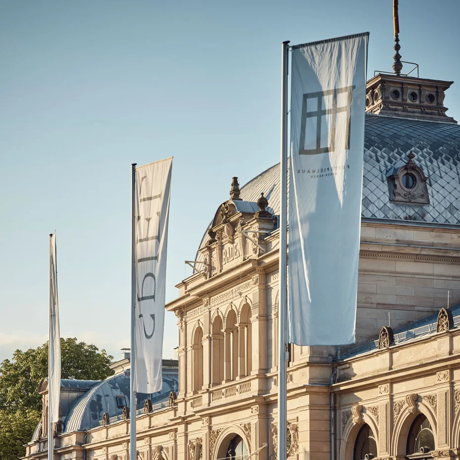 Festspielhaus Baden-Baden: Historisches Gebäude mit hoher Fassade und großen Fenstern. Drei lange Fahnen wehen vor dem Gebäude. Sonnenlicht beleuchtet die Szenerie.