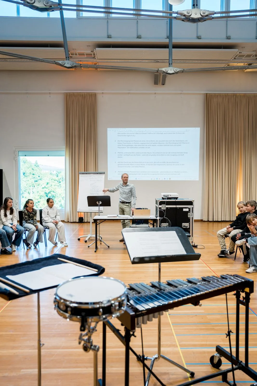 Ein Musikworkshop in einem großen Raum. Ein Vortragender spricht vor einer Projektionsleinwand. Instrumente sind im Vordergrund sichtbar.