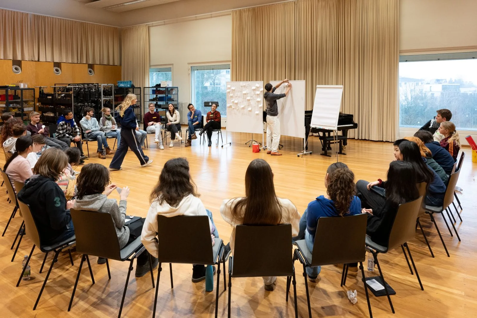 Ein Gruppenworkshop in einem Musiksaal. Menschen sitzen im Kreis, während jemand vor einer Tafel etwas erklärt.
