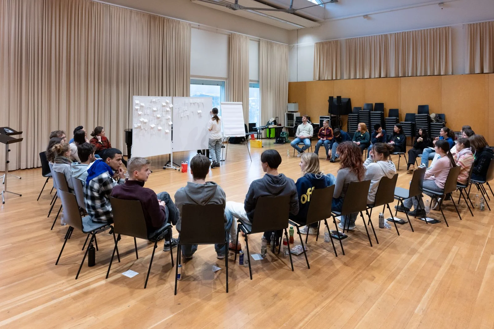 Ein Kreis von Menschen in einem Saal, die eine Präsentation oder ein Meeting verfolgen. Ein Moderator steht neben Tafeln.