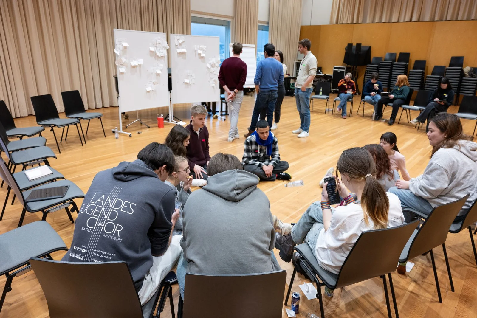 Eine Gruppe von Menschen sitzt in einem Raum und arbeitet in Teams. Im Hintergrund stehen Tafeln und weitere Personen.