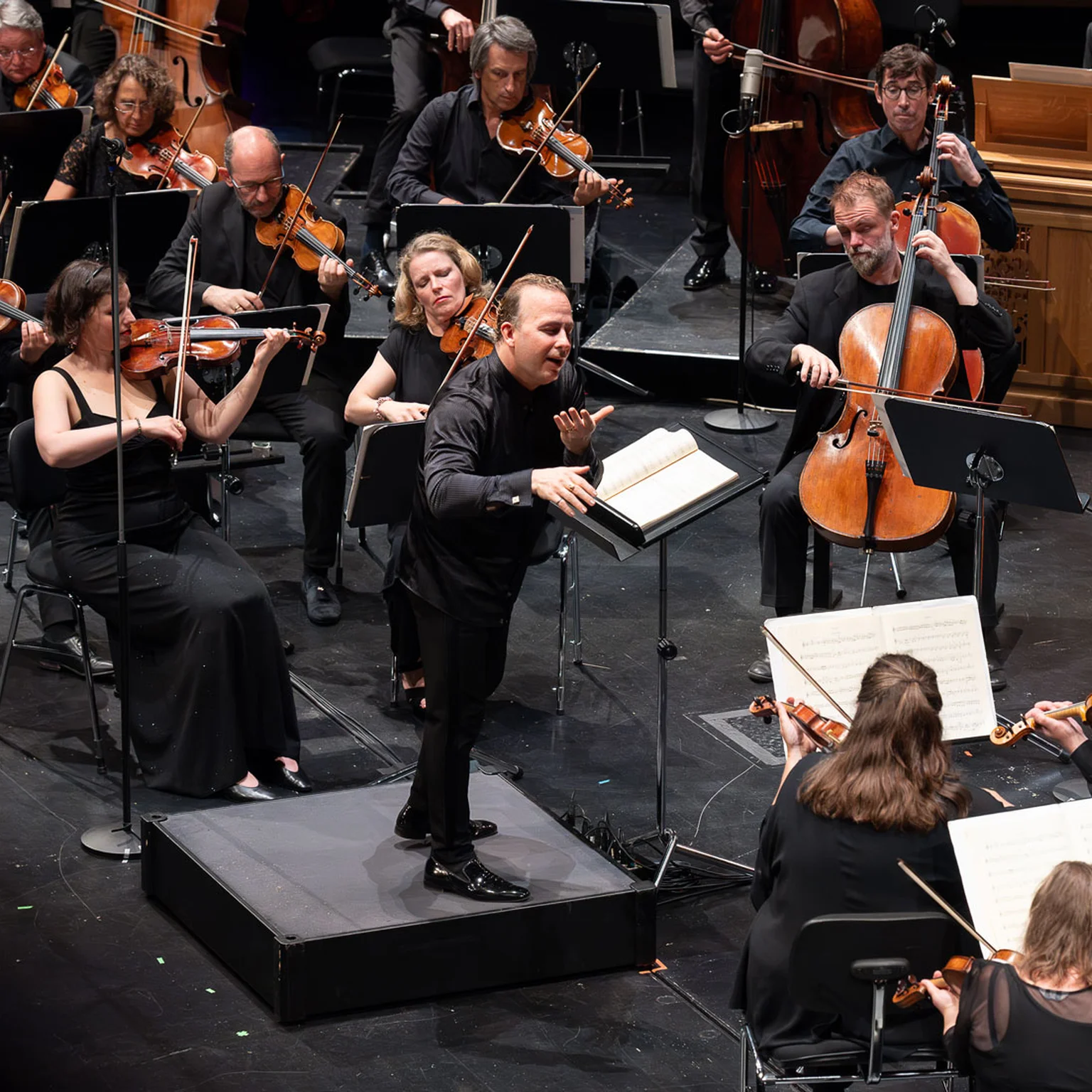 Ein Dirigent leitet ein Orchester auf einer Bühne. Musiker spielen verschiedene Streichinstrumente, während sie von Notenpulten lesen.