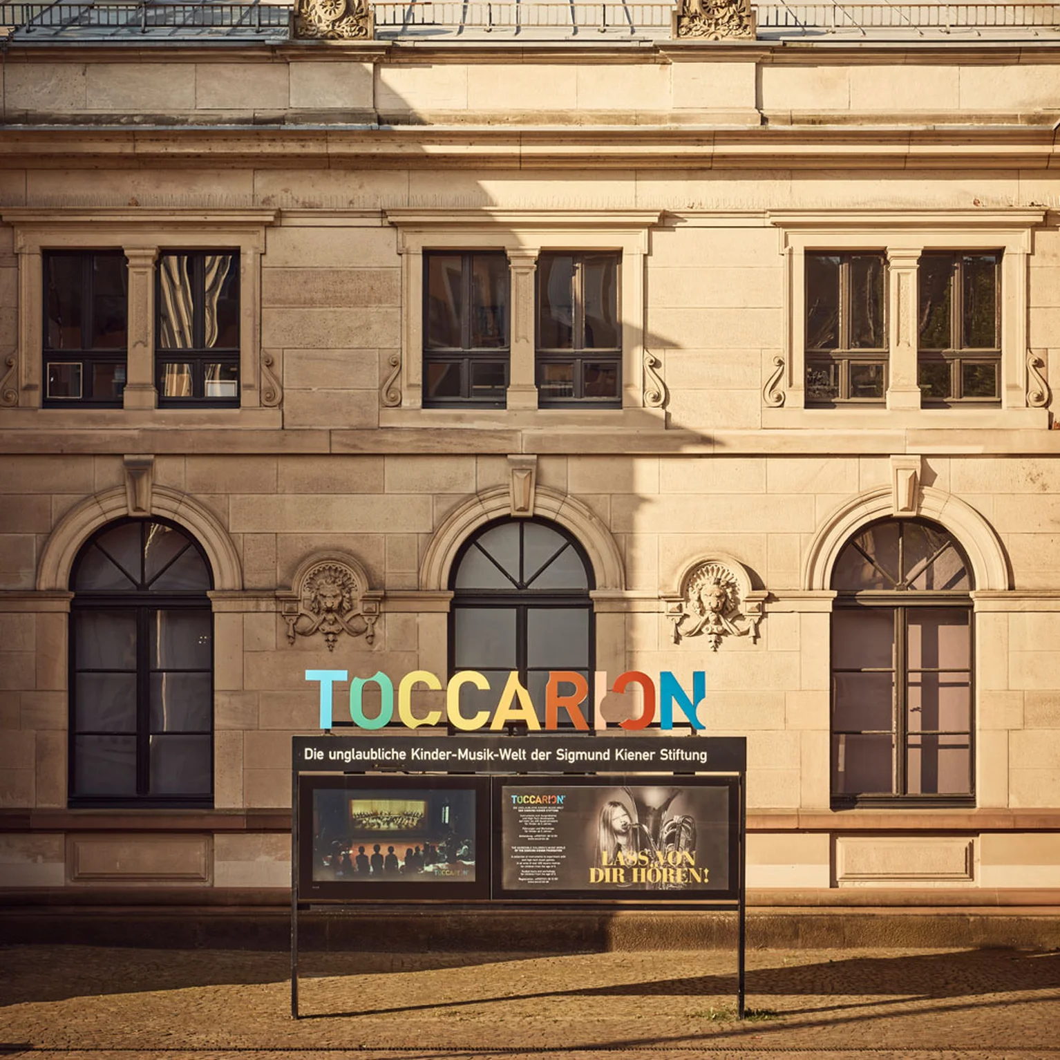 Außenansicht eines historischen Gebäudes mit einem farbigen Schild, auf dem "TOCCARION" steht. Moderne Architektur und Pflanzen im Hintergrund.