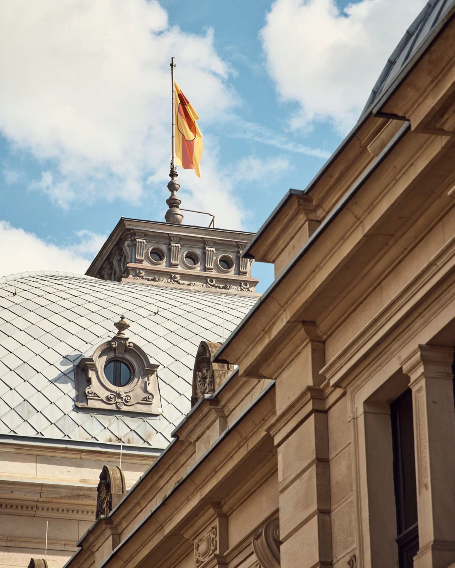 Architektonisches Detail eines historischen Gebäudes mit Flachdach und mehreren Stockwerken, mit einer Fahne auf dem Dach vor blauem Himmel.