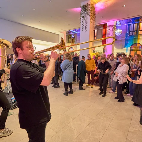 Musiker spielt Posaune bei einem lebhaften Event mit tanzenden und lachenden Menschen in einem farbenfroh beleuchteten Raum.