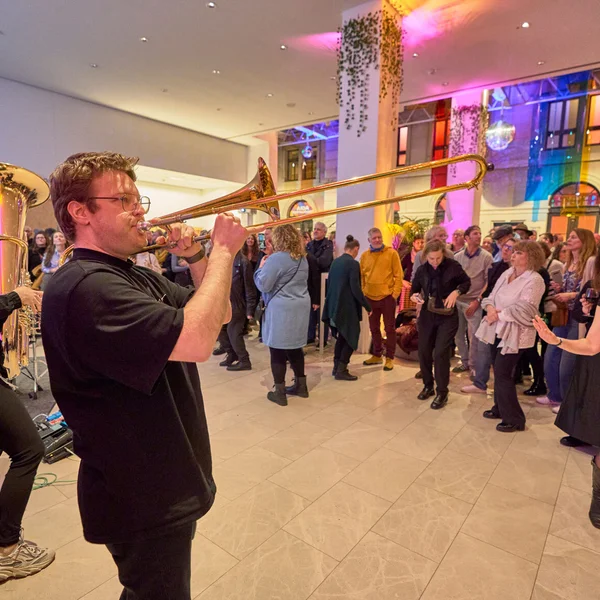 Musiker spielt Posaune bei einem lebhaften Event mit tanzenden und lachenden Menschen in einem farbenfroh beleuchteten Raum.