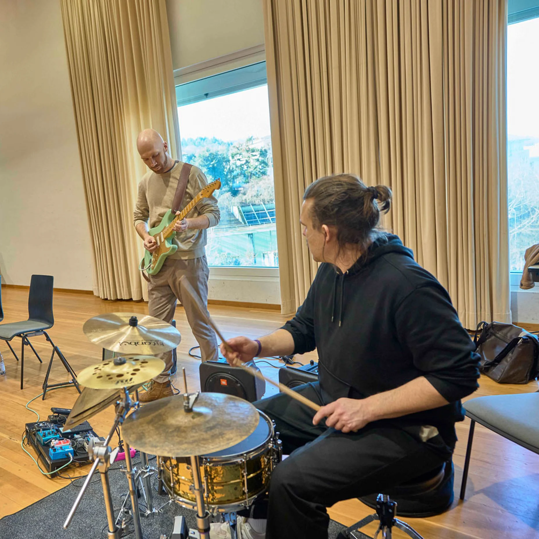Eine Band probt in einem Raum mit großen Fenstern. Ein Schlagzeuger, Gitarrist und zwei weitere Musiker sind zu sehen.