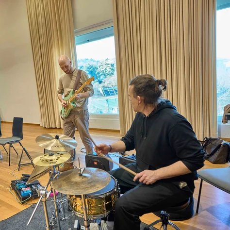Eine Band probt in einem Raum mit großen Fenstern. Ein Schlagzeuger, Gitarrist und zwei weitere Musiker sind zu sehen.